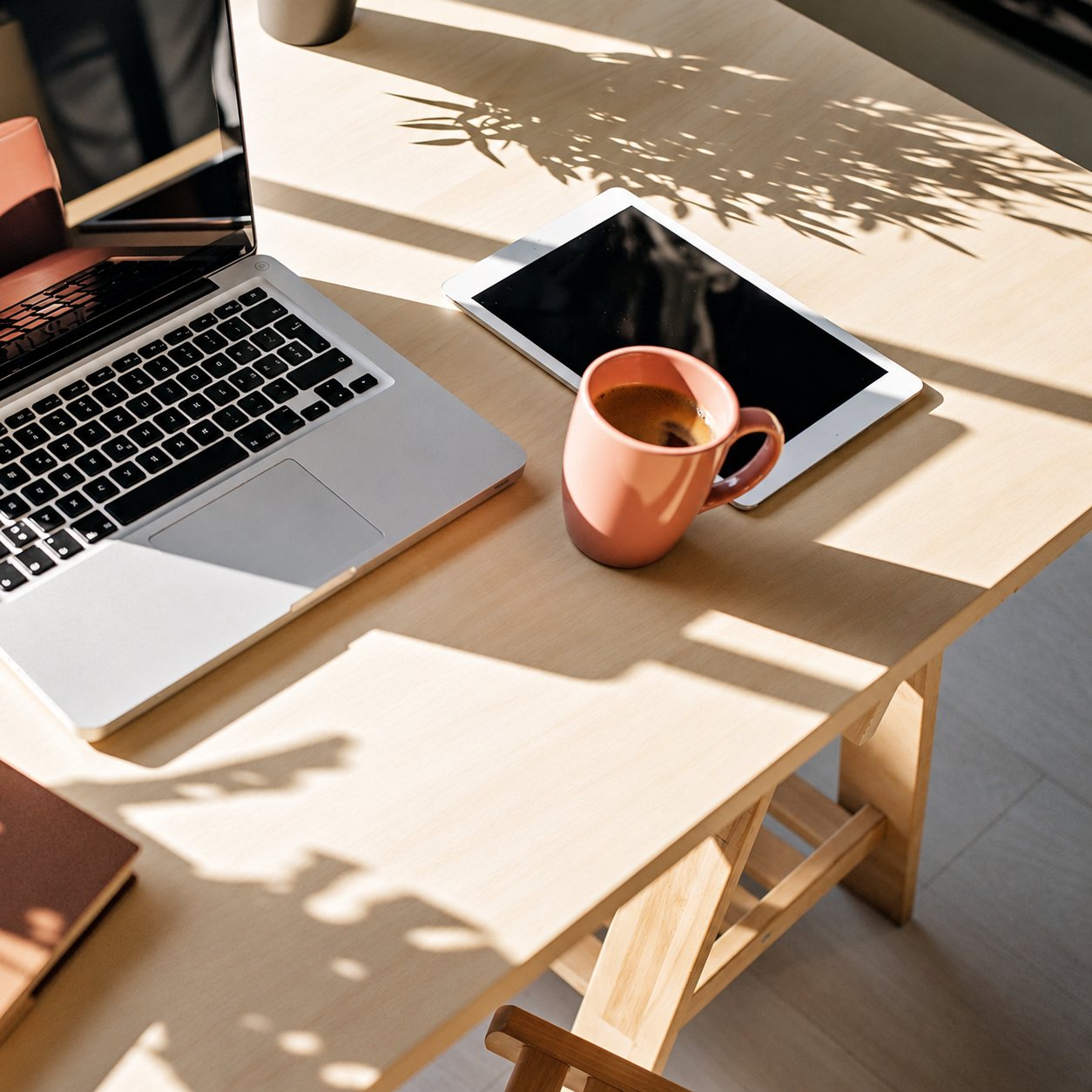 Un bureau en bois clair baigné de soleil avec un ordinateur portable, une tablette et une tasse de café.
